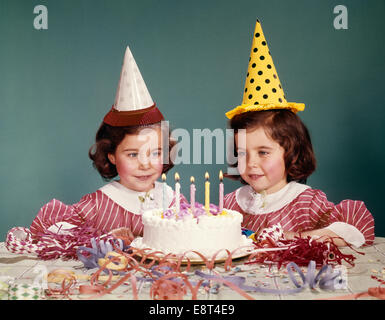 Anni sessanta figlie gemelle indossando cappelli di partito e la torta di compleanno con cinque candele Foto Stock