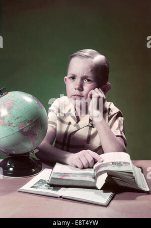 Negli anni quaranta anni cinquanta ragazzo seduto alla scrivania con globo la lettura di un libro di testo in appoggio mento a portata di mano Foto Stock