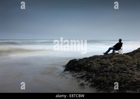 Uomo caucasico in cattedra si affaccia su onde sulla spiaggia rocciosa Foto Stock