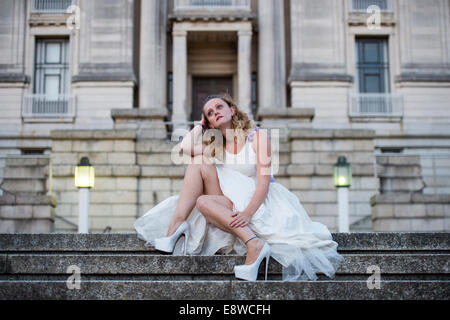 Il runaway o jilted Sposa - una giovane donna ragazza in un abito da sposa seduto da solo triste sulla scalinata di pietra, sera Foto Stock