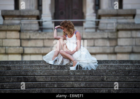 Il runaway o jilted Sposa - una giovane donna ragazza in un abito da sposa seduto da solo triste sulla scalinata di pietra, serata all'aperto Foto Stock