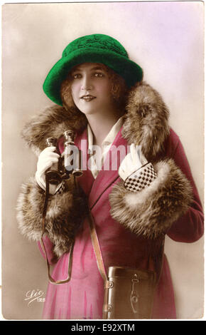 Moda Donna in Fur-Trimmed cappotto e cappello verde tenendo il binocolo Hand-Colored cartolina, Francia, circa 1922 Foto Stock