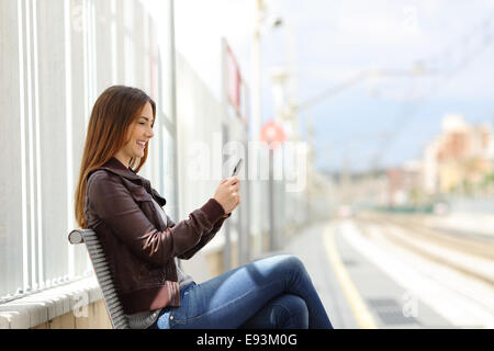 Donna felice scrivere messaggi su un telefono intelligente nella stazione ferroviaria con la stazione ferroviaria in background Foto Stock