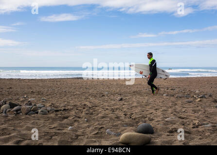 Giovane maschio camminare su una spiaggia di sabbia, portante una tavola da surf. Foto Stock