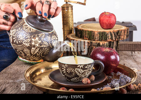 Donna versando da tè teiera su vintage tavolo in legno con nocciole e mele golden coffee mill in stile tradizionale Foto Stock