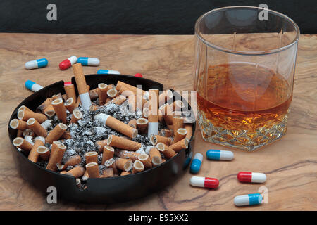 Sigari su un portacenere pieno di cenere, pillole e whiskey su un tavolo di legno Foto Stock