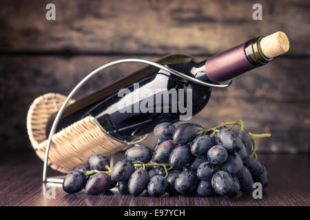 Cantina dello sfondo. Bottiglia di vino rosso nel supporto con il cluster di chicchi di colore blu scuro su un tavolo di legno. Immagine dai toni Foto Stock