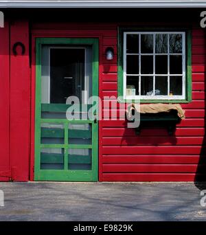 Vista di colore rosso brillante fienile muro con un bianco luminoso con finestra incorniciata e luminosa porta verde in una giornata di sole. Foto Stock