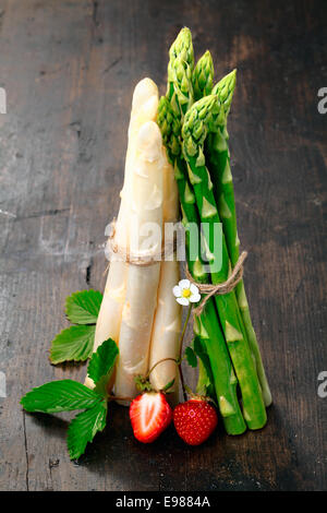 Montante grappoli di fresco verde e asparagi bianchi spears equilibrata su un tavolo di legno top guarnito con fragole Foto Stock