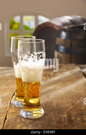 Due metà bevuto bicchieri di birra con una buona testa di schiumato in piedi su un vecchio rustico tavolo in legno con vintage botti di legno in background Foto Stock