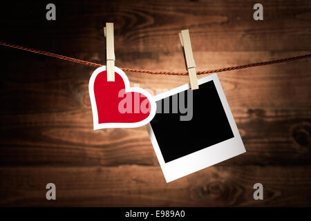 Un rosso e bianco cuore pende da una clothespeg in legno a fianco di uno sbozzato instant photo frame su un legno rustico sfondo per il vostro romantico nostalgici ricordi o messaggio di San Valentino Foto Stock