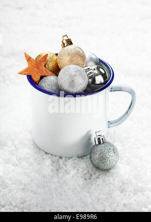 Le decorazioni di Natale in un stagno smaltato mug sulla neve che è pieno di traboccante di baubles e stelle Foto Stock