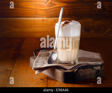 Stratificato delizioso cappuccino o latte macchiato caffè servito in un bicchiere a lungo con un cucchiaio contro un rustico in legno con sfondo copyspace Foto Stock