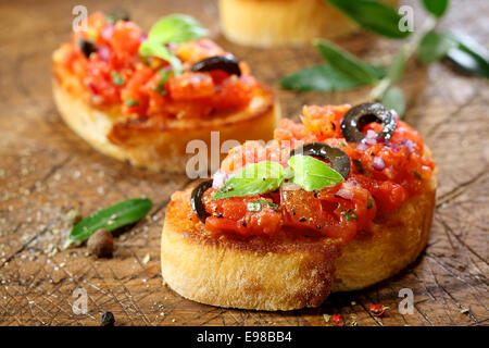 Preparare deliziose bruschette di pomodoro con un trito di verdure, erbe e olio su grigliati o tostate baguette cosparsi di condimenti e spezie su un vecchio grungy tagliere di legno Foto Stock