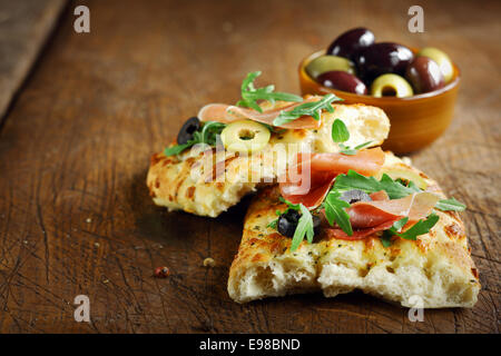 Prosciutto e rucola fresca sulla focaccia italiana guarnita con fette di olive giacente su una vecchia cucina in legno tavolo Foto Stock