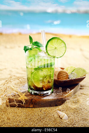 Caipirinha Rum e cocktail di calce guarnite con foglioline di menta e servita su una tavola di legno sulla sabbia dorata di una spiaggia tropicale Foto Stock