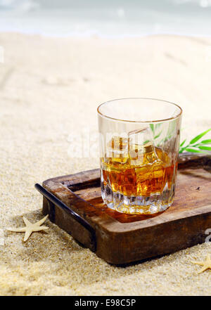 Bella fotografia di un bicchiere di whisky con cubetti di ghiaccio in un elegante vassoio in legno sulla spiaggia. Guardate il mio portafoglio per tutta una serie di cocktail. Foto Stock