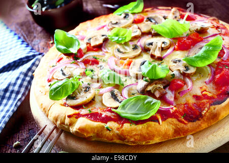 Una deliziosa pizza vegetariana condito con pomodoro, formaggio fuso, funghi, cipolle e di foglie di basilico fresco servito su una tavola di legno, ad alto angolo di visione. Più la pizza alla mia porta. Foto Stock