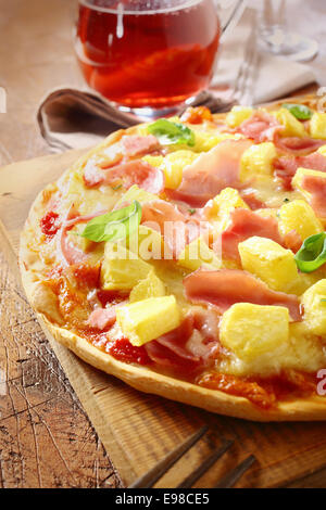 Vista ingrandita di un appetitoso prosciutto e ananas pizza italiana con un croccante crosta dorata servita su un vecchio grunge tavolo in legno con un bicchiere di vino rosso Foto Stock