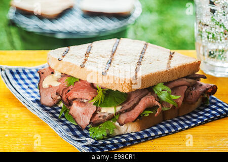 Grigliata di sandwich tostato con rare roast beef, rucola e erbe su un blu e bianco panno controllato all'aperto su un tavolo da picnic a Foto Stock