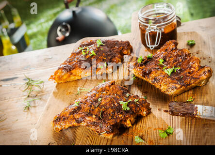 Inumidendo di stagionatura e nervature per il barbecue con tre porzioni di carne steso su una tavola di legno tavolo da picnic in un giardino estivo con un barattolo di salsa di accompagnamento ed erbe aromatiche Foto Stock