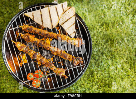 Vista dall'alto di due di agnello, manzo o spiedini di carne di maiale con fette di pane tostato e pomodori grigliare su un barbecue il fuoco su un portatile barbecue di metallo durante un picnic estivo su erba verde con copyspace Foto Stock