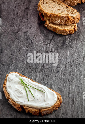Saporito formaggio spalmabile su una fetta di pane di segale guarnita con erba cipollina fresca con ulteriori fette di pane formando un angolo diagonale sul confine di una texture di sfondo scuro con copyspace Foto Stock