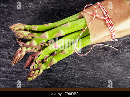 Fresco verde i turioni degli asparagi legate in un fascio con carta marrone e string, vicino la vista aerea su ardesia scura con copyspace Foto Stock