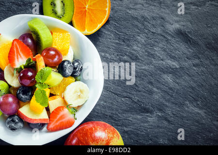 Vista aerea di una ciotola di insalata di frutta fresca per una dieta sana fatta con un assortimento di frutta tropicale e una mela, kiwi e arancio disposte affiancate su ardesia testurizzata con copyspace Foto Stock