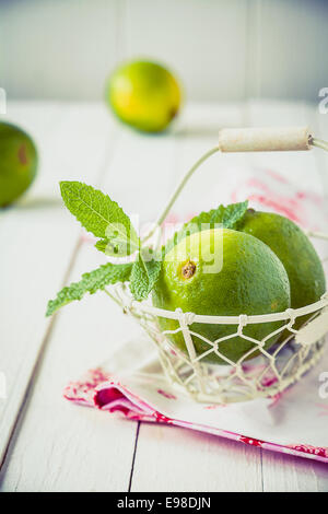 Piccolo cestino con lime e foglie di menta su un tovagliolo e un rustico di legno bianco tabella Foto Stock