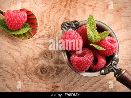 Vista dall'alto di un vecchio mestolo di rame e conchiglia forma di cucchiaio riempito con lamponi maturi decorato con foglie di menta su un tavolo di legno Foto Stock