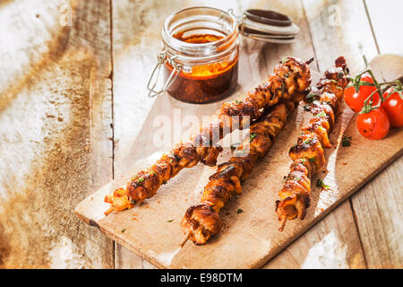 Piccante di carne alla brace spiedini su un picnic estivo servita su un tagliere di legno con pomodori arrosto al fianco di un vasetto di piccante salsa di accompagnamento, con copyspace Foto Stock