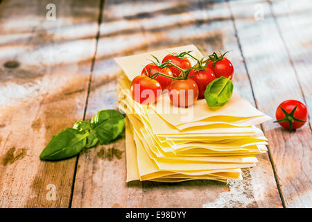 Ingredienti per le lasagne italiane con freschi pomodori ciliegia e il verde del basilico foglie su fogli di pasta essiccata su un legno rustico tavolo da cucina Foto Stock