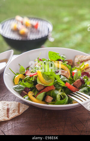 Fresche erbe sane con insalata di rucola e erbe miste con peperone dolce e pomodoro su un tavolo da picnic con un barbecue estivo sul prato dietro Foto Stock