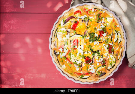 Appena sfornato quiche salate con melanzane, peperoni, pomodori e formaggio stagionato con erbe fresche nelle pieghe di una teglia, vista aerea con copyspace su rosso di tavole di legno Foto Stock