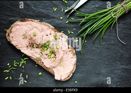 La preparazione di un saporito affumicato sandwich teewurst su una fetta di pane di segale con erba cipollina tritata e pepe nero, vista aerea su una cucina in ardesia con contatore copyspace Foto Stock