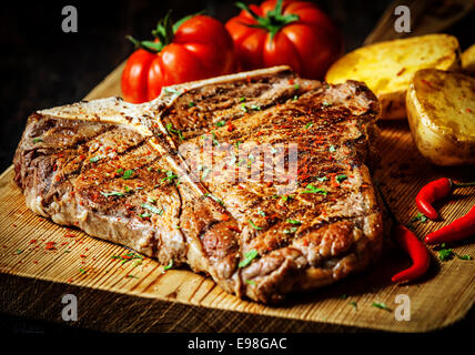 Grigliata di T-bone steak conditi con spezie ed erbe fresche servita su una tavola di legno con pomodoro fresco , patate arrosto e red hot chili peppers Foto Stock