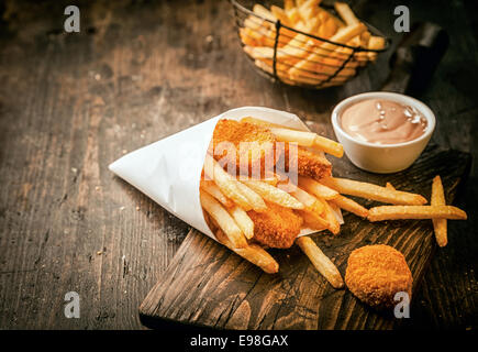 Servire in un cono di carta di takeaway impanati fritti pepite di pesce con patatine e una piccola ciotola di salsa o dip su una tavola in legno rustico con copyspace Foto Stock