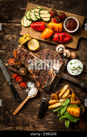 Ricca la diffusione con t-bone o osteria bistecca servita con un assortimento di sano verdure arrosto e cali di salato su una tavola in legno rustico in un paese cucina Foto Stock