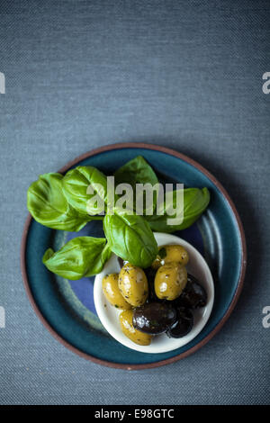 Vista aerea di un mazzo di foglie di basilico fresco con una ciotola di nero e verde olive indurita su un intreccio di grigio Sfondo tessile con copyspace Foto Stock