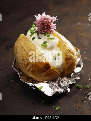 Grigliata di patate al forno sul suo involucro in foglio servita con un ripieno di panna acida, tritato fresco verde erba cipollina e un fiore di erba cipollina su sfondo scuro Foto Stock