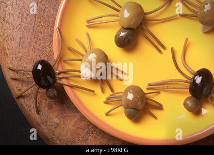 Carino spooky Halloween spider antipasti dal verde, nero e Olive ripiene con spaghetti gambe strisciando fuori da un piatto giallo su un tavolo di legno, vista da sopra Foto Stock