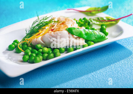 Serve della deliziosa cotta al forno o alla brace filetto di pesce con piselli , baby spinaci foglie e la scorza di limone guarnito con aneto su un piatto bianco, sfondo blu Foto Stock