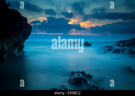 Nuvoloso Tramonto su scogli lungo la costa della Cornovaglia vicino a Newquay, Cornwall, Inghilterra Foto Stock