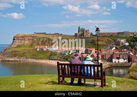 Due persone anziane coppia seduta sulla panchina che si affaccia sul Abbazia e porto in estate Whitby North Yorkshire Inghilterra UK Regno Unito Gran Bretagna Foto Stock