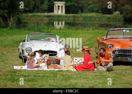 Famiglia avente un picnic nella parte anteriore di una coppia di auto classiche con un piccolo lago e un tempio greco follia dietro Foto Stock