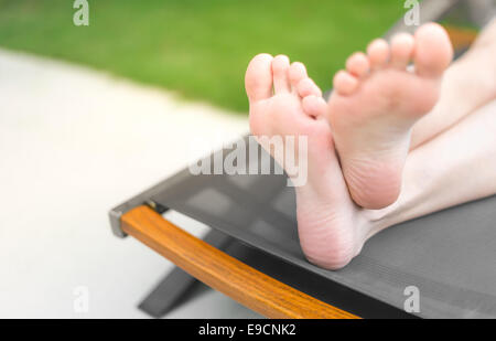 Focus su piedi incrociati di persona sdraiata e abbronzatura sul lettino grigio. Prato verde in background. La donna per le gambe con la pelle liscia. Foto Stock