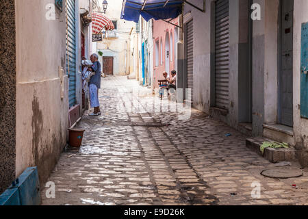 Un uomo raccoglie biancheria da una casa giù per una strada laterale nel cuore della medina a Sousse, Tunisia mentre altri due chat su una porta. Foto Stock