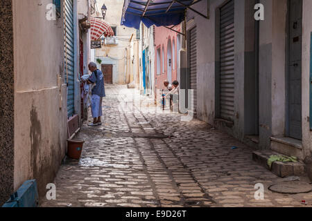 Un uomo raccoglie biancheria da una casa giù per una strada laterale nel cuore della medina a Sousse, Tunisia mentre altri due chat su una porta. Foto Stock