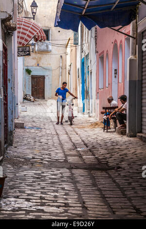 Giovane uomo cammina il suo ciclomotore giù una stretta e acciottolata strada laterale nella Medina di Sousse,Tunisia mentre altri due chat su una porta. Foto Stock